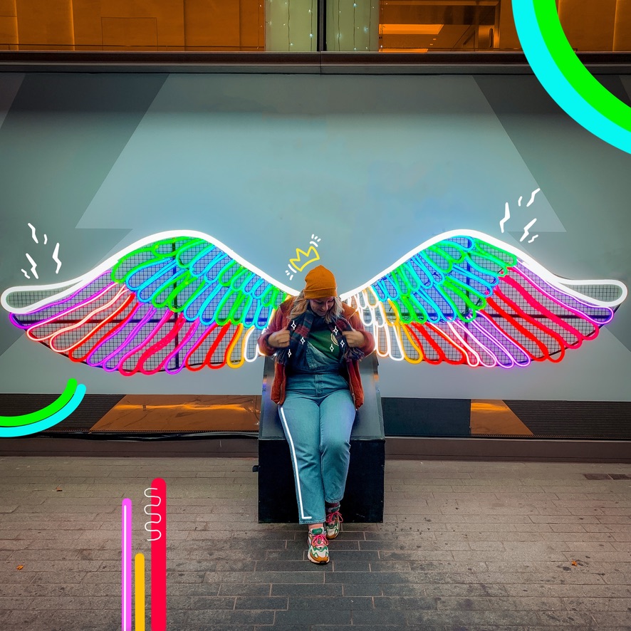 Wendy standing in front of a statue with neon wings in different colors. She's standing in front of it, making it look like the wings are hers. On the photo there are colorful shapes drawn and a little crown on her head.