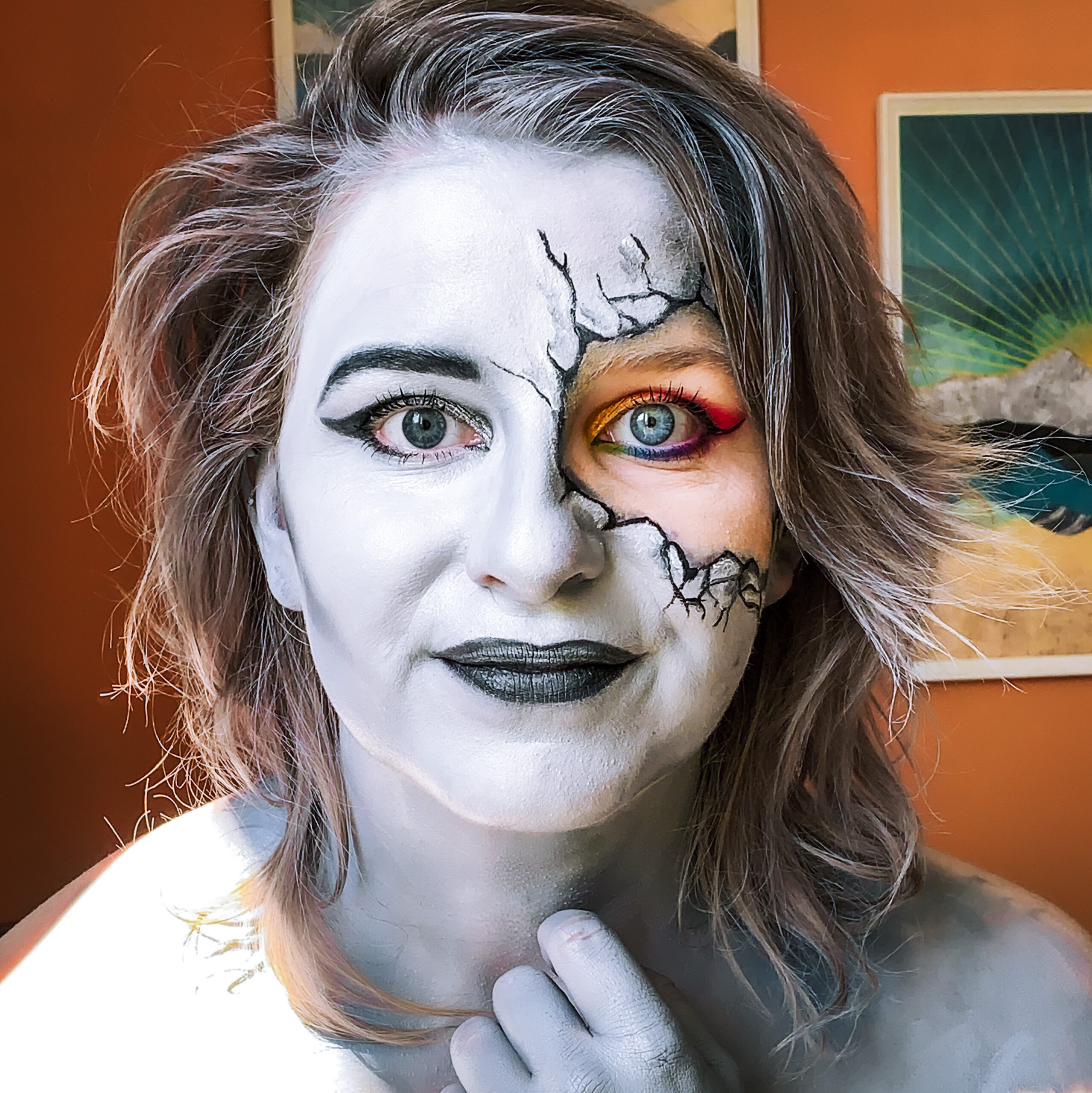 Wendy with black/white/grey make up on, except around one of her eyes, there we see colorful makeup coming through cracks drawn around the eye. The background is orange and Wendy stares directly at the camera.