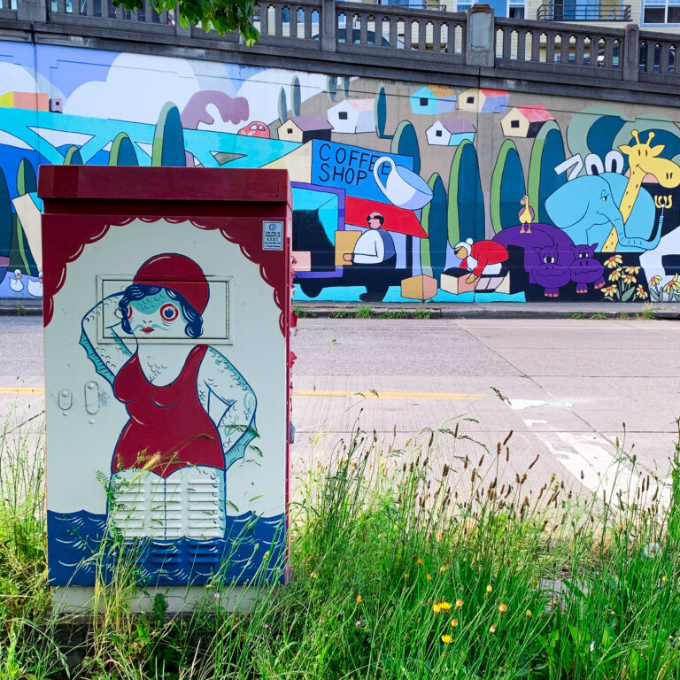 An electricity box with a drawing of a fish person on it. They have a mustache, breasts and are wearing swimming gear. In the background is colorful grafitti.