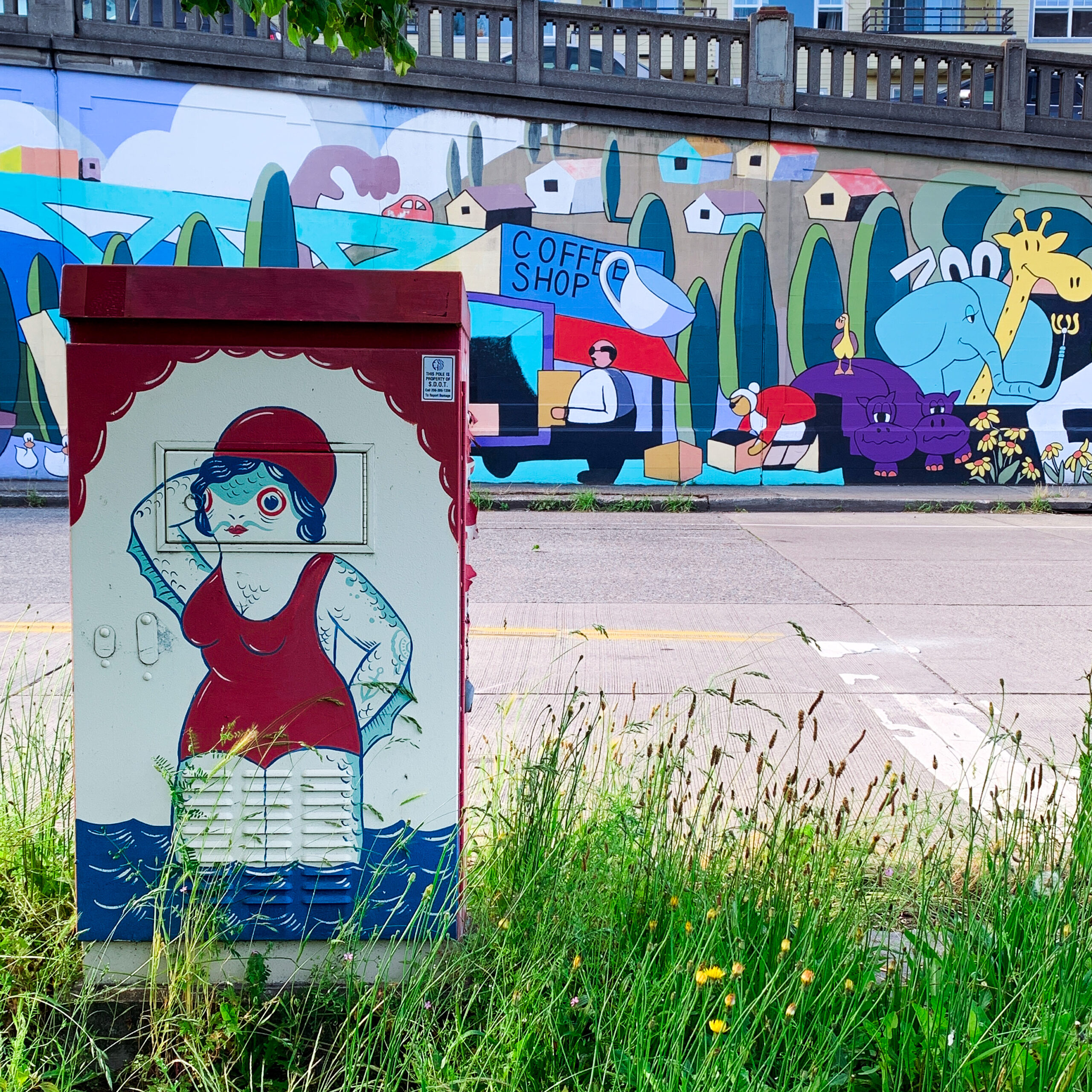 An electricity box with a drawing of a fish person on it. They have a mustache, breasts and are wearing swimming gear. In the background is colorful grafitti.