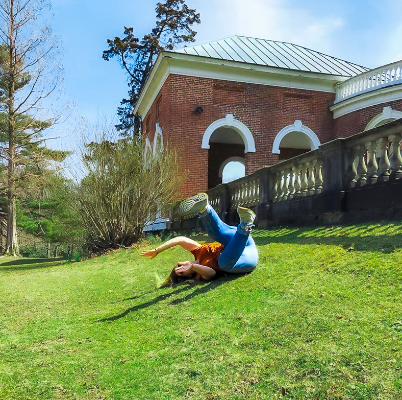 Wendy rolling down a slope on a grass field next to a building with a little wall.