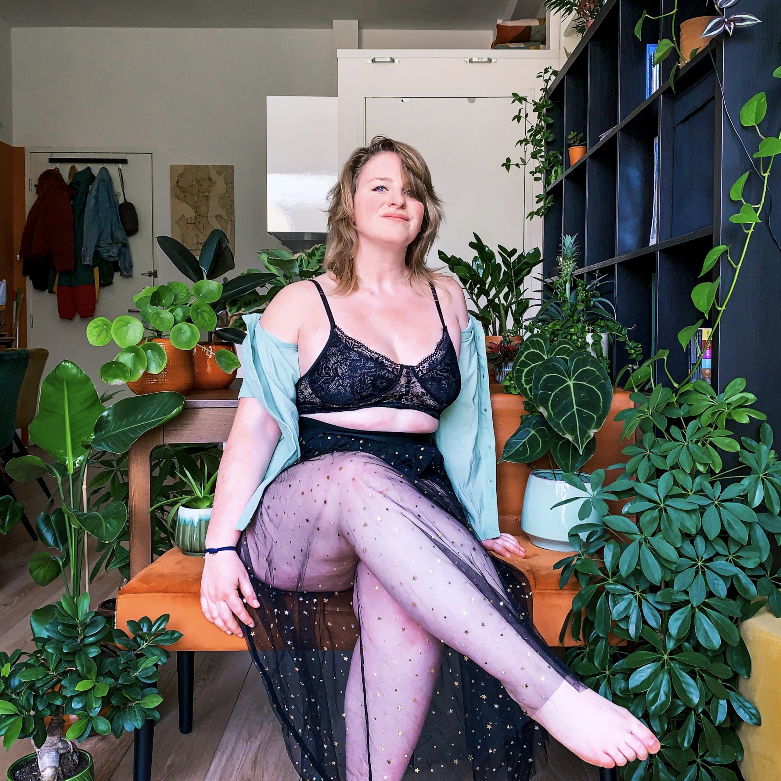 Wendy in a skirt and bra, looking at the camera while sitting in front of plants.