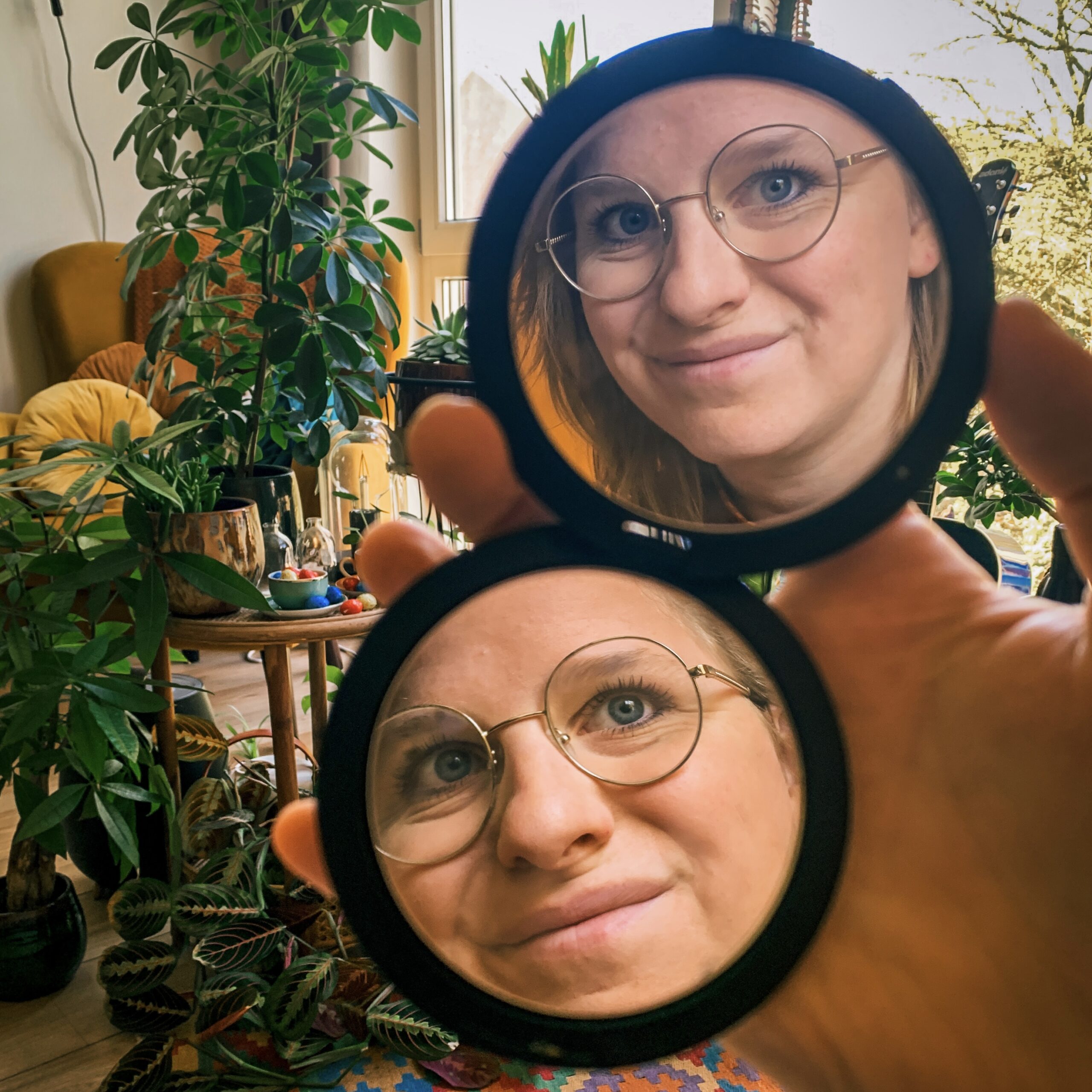 Wendy looking into a compact mirror with two sides. On one side she looks straigt at the camera, on the other side she looks into the distance. In the backgrounds we can see plants.