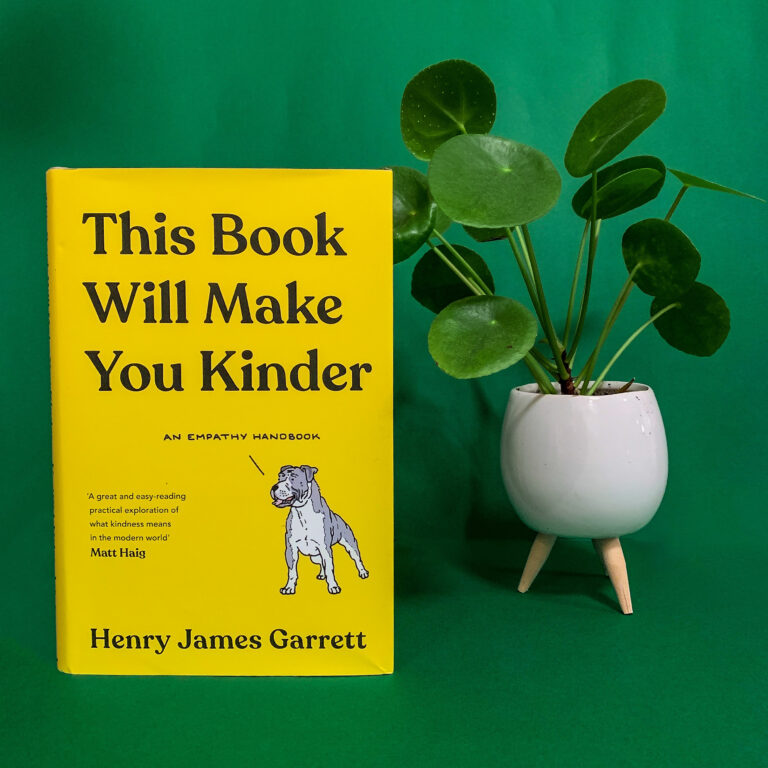 A photo of the book 'This Book Will Make You Kinder' by Henry James Garrett in front of a green backdrop and next to a plant